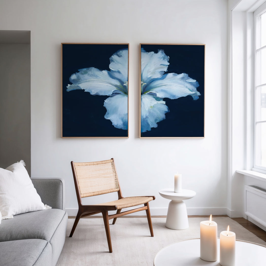 A modern living room features a light gray sofa, woven chair, and round white side table with candles. Two Luminous Iris - Set of 2 canvas prints with blue backgrounds hang above the chair as natural light streams through tall windows.