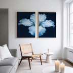 A modern living room features a light gray sofa, woven chair, and round white side table with candles. Two Luminous Iris - Set of 2 canvas prints with blue backgrounds hang above the chair as natural light streams through tall windows.