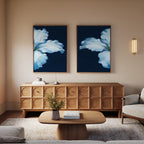 A modern living room features a wooden sideboard, Luminous Iris - Set of 2 canvas prints with dark blue backgrounds, a round coffee table, beige armchair, wall sconce, and a vase with flowers on the table.