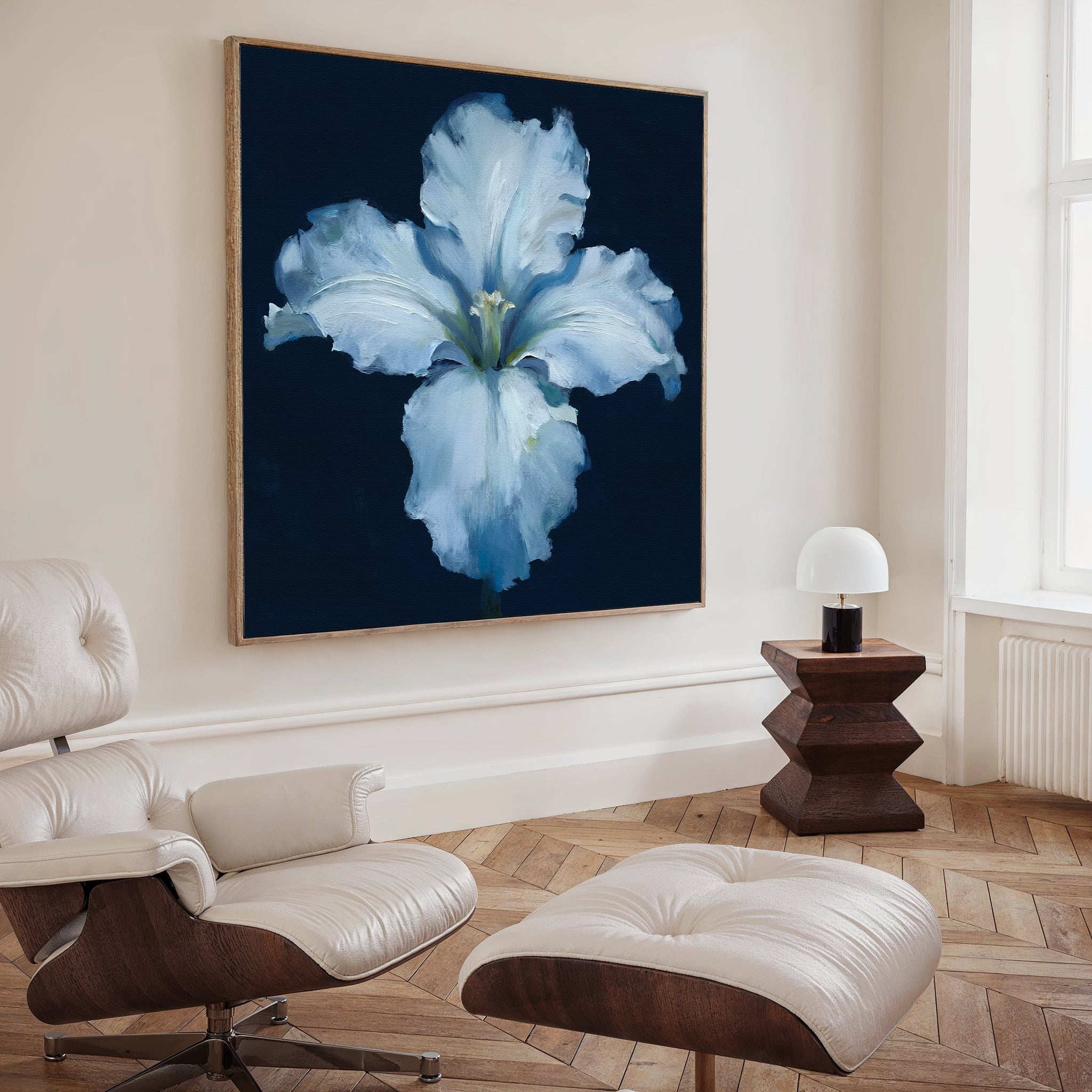 A modern living room with a white lounge chair and ottoman, wooden side table and lamp, and the Luminous Iris Canvas Art - Square in blue and white from Nordic Park displayed on the wall.
