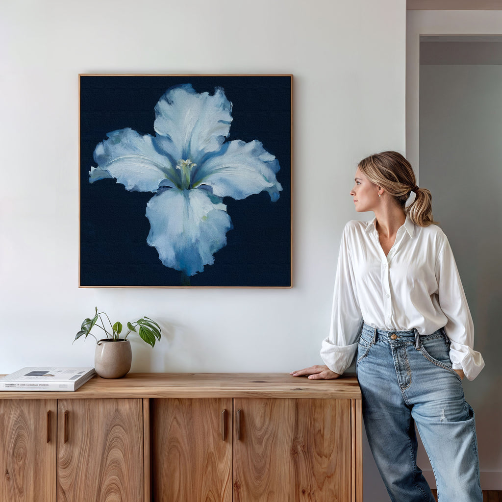 A woman in a white blouse and jeans leans against a wooden cabinet, admiring the Luminous Iris Canvas Art - Square on the wall above—a radiant flower piece that adds Nordic elegance to the space.