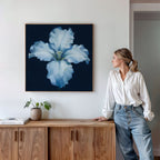 A woman in a white blouse and jeans leans against a wooden cabinet, admiring the Luminous Iris Canvas Art - Square on the wall above—a radiant flower piece that adds Nordic elegance to the space.