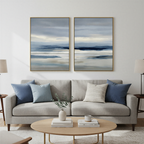 A modern living room with a light gray sofa, blue and patterned cushions, a round wooden coffee table with a plant, and the Blue Mist Horizon Set of 2 abstract landscape paintings above the sofa.