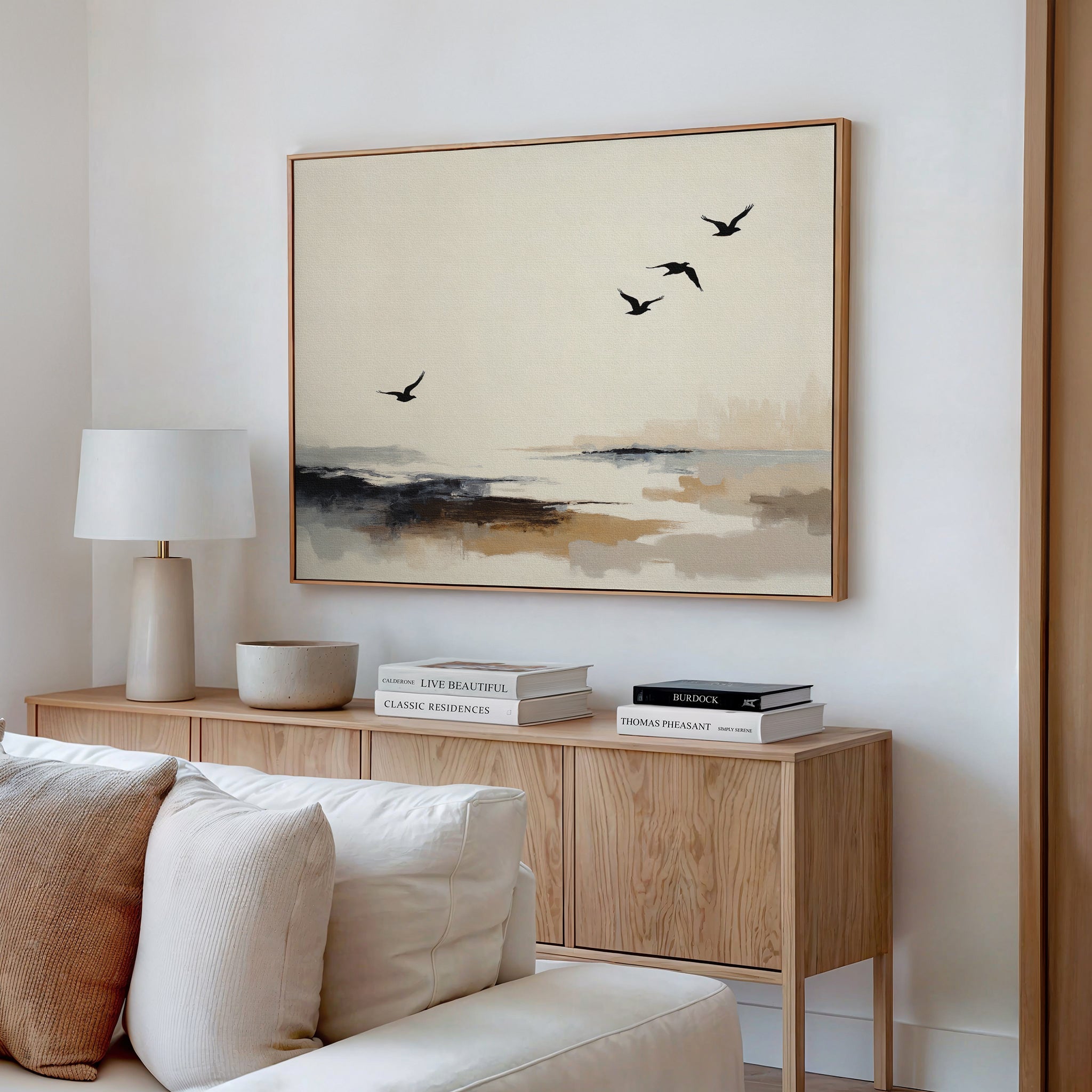 A modern living room with a light wood sideboard, stacked books, a lamp, and neutral decor. Above the sideboard hangs the Maritime Flight Canvas Art - Horizontal, featuring four birds over an abstract landscape in soft beige and gray tones.