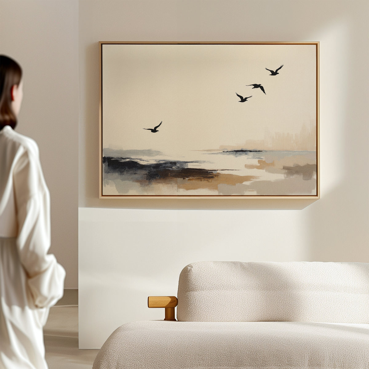 In a minimalistic room, a person admires the Maritime Flight Canvas Art - Horizontal, showcasing four birds soaring over a serene abstract landscape in neutral tones above a light-colored sofa.