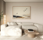 A modern, minimalist living room features a light beige sectional sofa, a low wooden coffee table with a black bowl, and the Maritime Flight Canvas Art - Horizontal depicting birds over an abstract landscape. Neutral tones and soft lighting add warmth.
