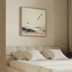 A beige bedroom with a cushioned headboard, white bedding, and four pillows. Above the bed hangs the Maritime Flight Canvas Art - Square, depicting flying birds in coastal tones. A vase with branches sits on a bedside table.