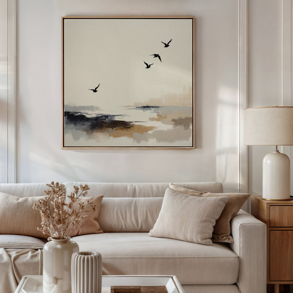 A modern living room with a beige sofa, neutral cushions, and a side table with a ceramic lamp. Above the sofa hangs the Maritime Flight Canvas Art - Square, featuring four birds over an abstract landscape. Dried flowers in a vase sit on the coffee table.