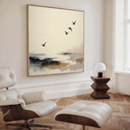 A modern, minimalist living room features a white lounge chair, ottoman, geometric wooden side table, small white lamp, and the Maritime Flight Canvas Art - Square, adding subtle nautical decor with birds soaring over an abstract coast.