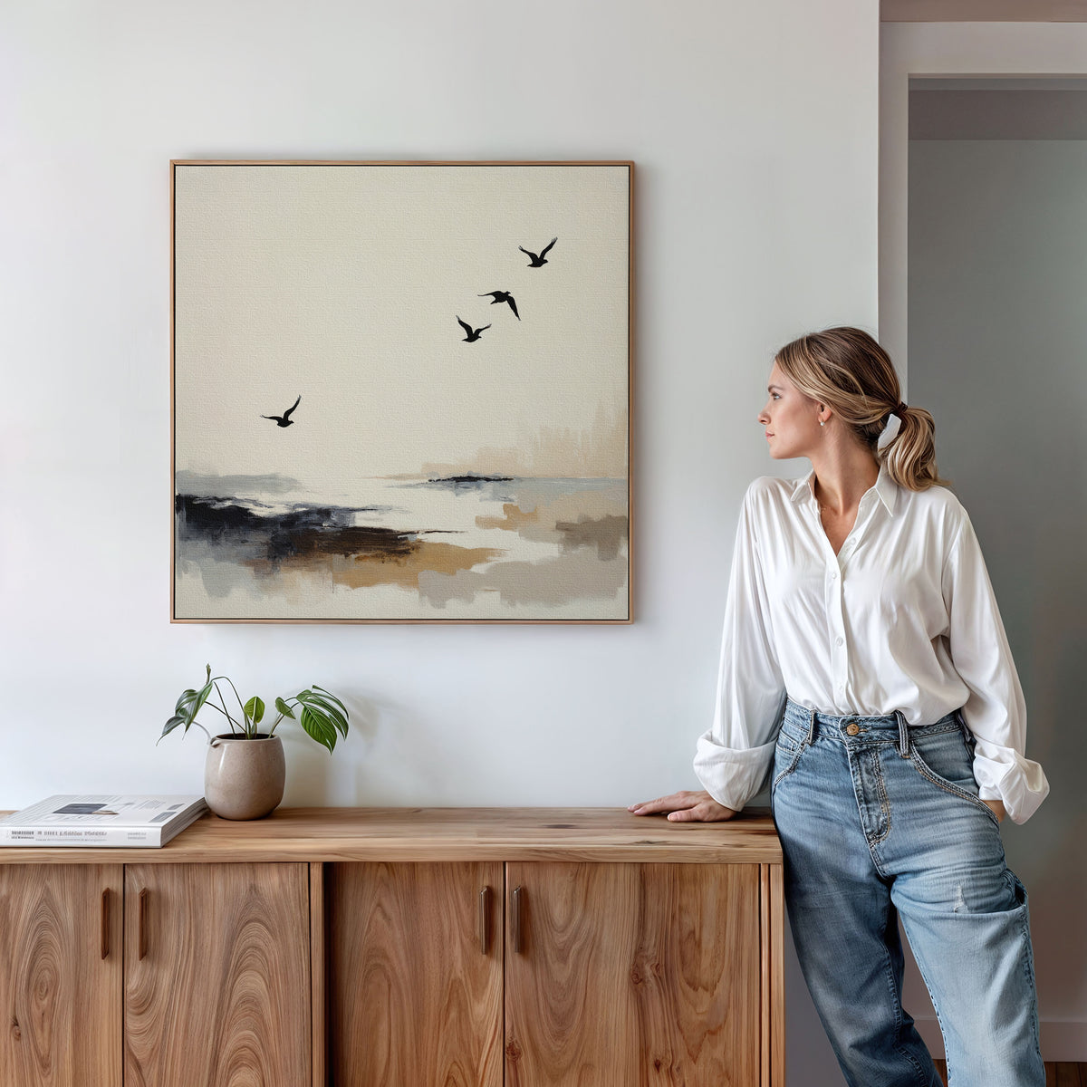 A woman in a white blouse and jeans leans on a wooden sideboard, admiring the Maritime Flight Canvas Art - Square, which depicts flying birds over a serene landscape. A small plant and books sit on the sideboard in this minimal, modern room.
