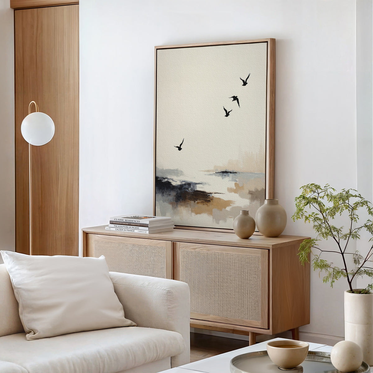 Minimalist living room with a light wood sideboard, coastal decor accents, potted plant, and the Maritime Flight Canvas Art - Vertical featuring abstract landscapes and flying birds. Light walls and a cozy white sofa complete the serene space.
