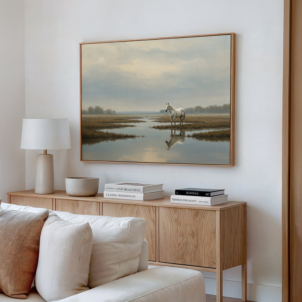 A modern living room features a light wood sideboard with books and a lamp, neutral decor, and the Marsh Reflection Canvas Art - Horizontal above, showing a Nordic Park landscape of a white horse standing in water.