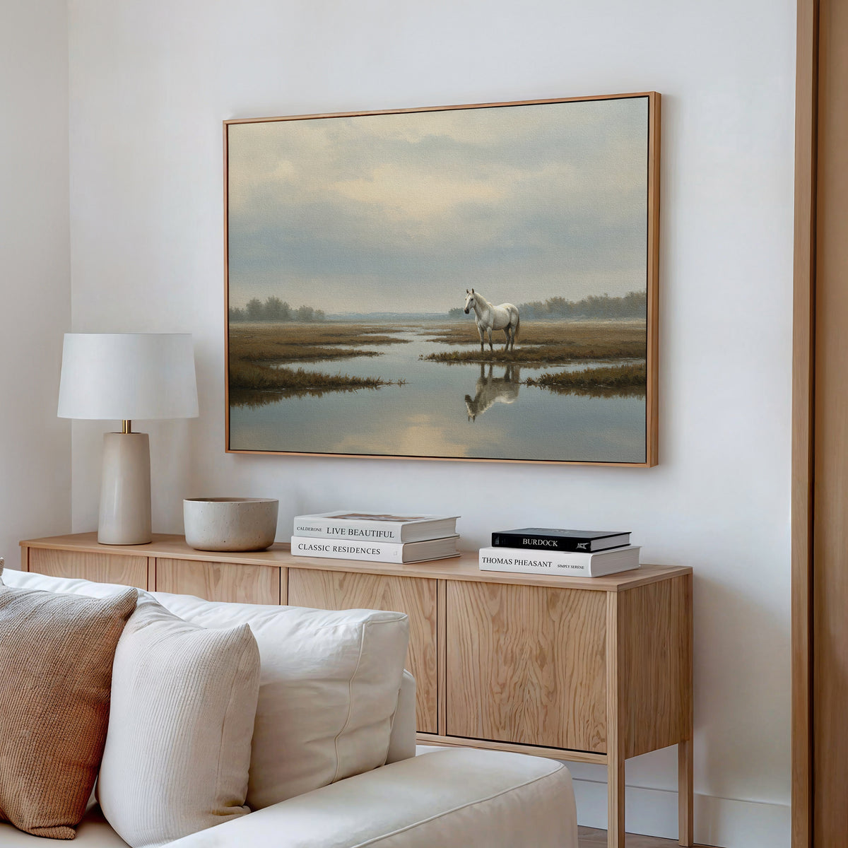 A modern living room features a light wood sideboard with books and a lamp, neutral decor, and the Marsh Reflection Canvas Art - Horizontal above, showing a Nordic Park landscape of a white horse standing in water.