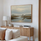 A modern living room features a light wood sideboard with books and a lamp, neutral decor, and the Marsh Reflection Canvas Art - Horizontal above, showing a Nordic Park landscape of a white horse standing in water.