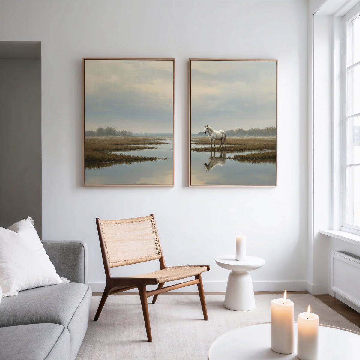 A modern, minimal living room with a gray sofa, wooden chair, white round side table, and candles. Above the chair, two framed Marsh Reflection - Set Of 2 prints of serene marshland landscapes hang on the white wall.