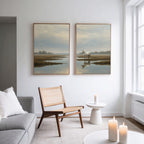 A modern, minimal living room with a gray sofa, wooden chair, white round side table, and candles. Above the chair, two framed Marsh Reflection - Set Of 2 prints of serene marshland landscapes hang on the white wall.
