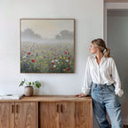 A woman in a white shirt and jeans stands by a wooden sideboard, admiring the Meadow Dreams Canvas Art - Square, featuring a misty meadow scene on a white wall. A potted plant and books sit on the sideboard.