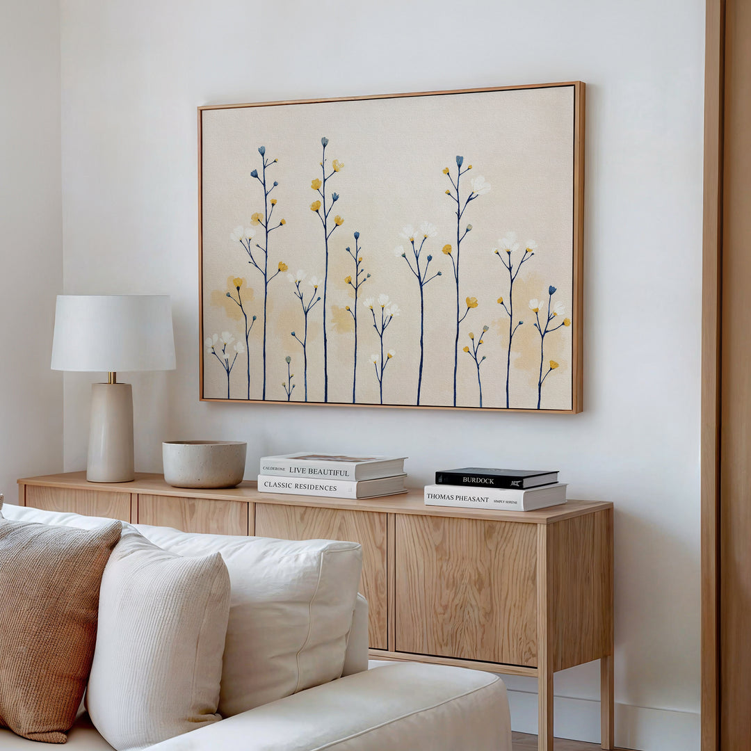 A modern living room features a wooden sideboard with books, a lamp, and a bowl. Above, the Meadow Symphony Canvas Art - Horizontal adds soft yellow and blue spring meadow tones to the white wall, with a white sofa partly visible in front.
