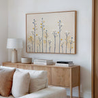 A modern living room features a wooden sideboard with books, a lamp, and a bowl. Above, the Meadow Symphony Canvas Art - Horizontal adds soft yellow and blue spring meadow tones to the white wall, with a white sofa partly visible in front.