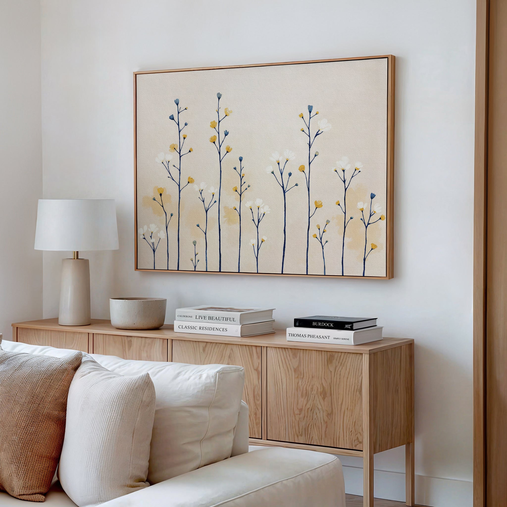 A modern living room features a wooden sideboard with books, a lamp, and a bowl. Above, the Meadow Symphony Canvas Art - Horizontal adds soft yellow and blue spring meadow tones to the white wall, with a white sofa partly visible in front.