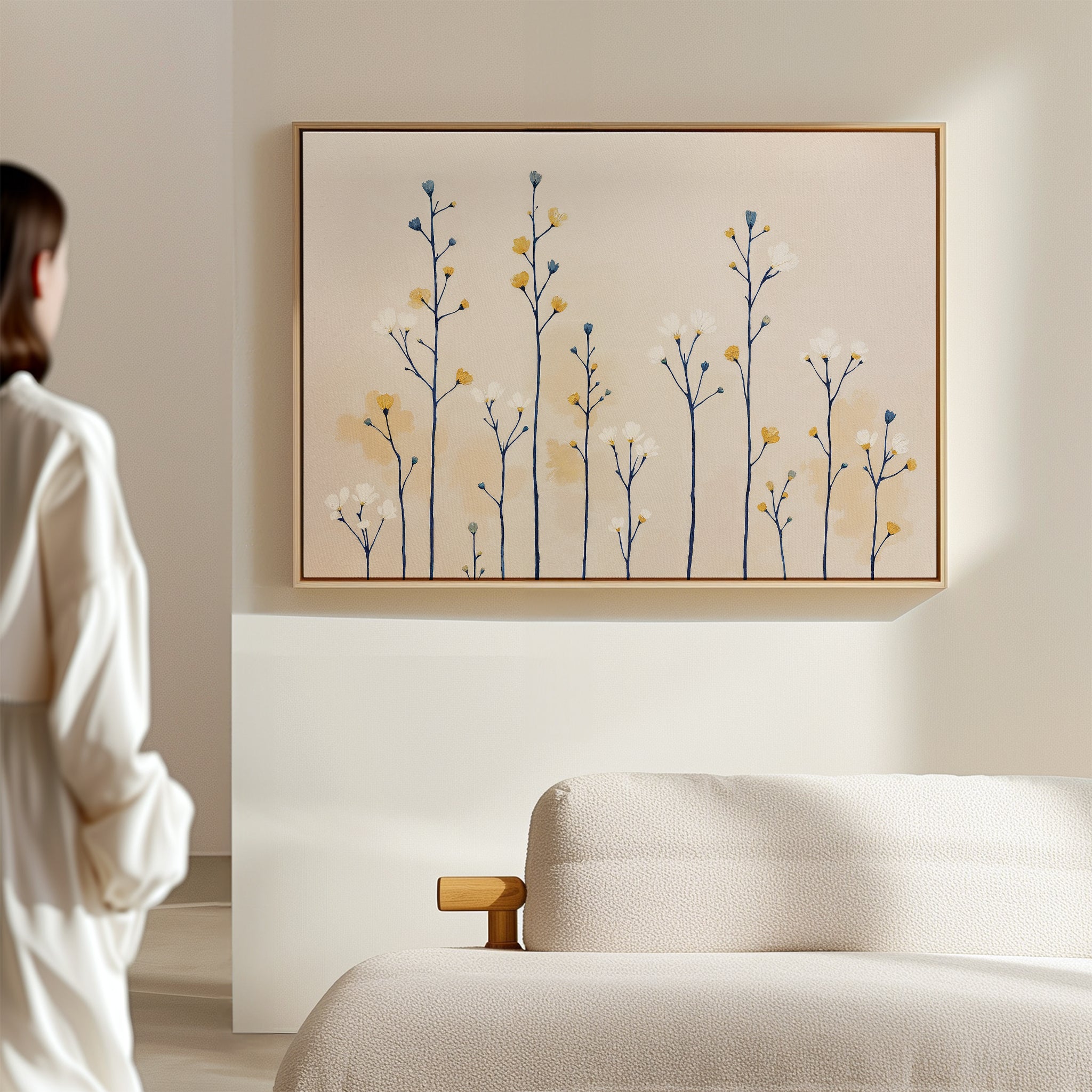 A person in a white shirt stands in a minimalistic room, gazing at the Meadow Symphony Canvas Art - Horizontal featuring delicate abstract yellow, blue, and white flowers above a beige textured sofa.
