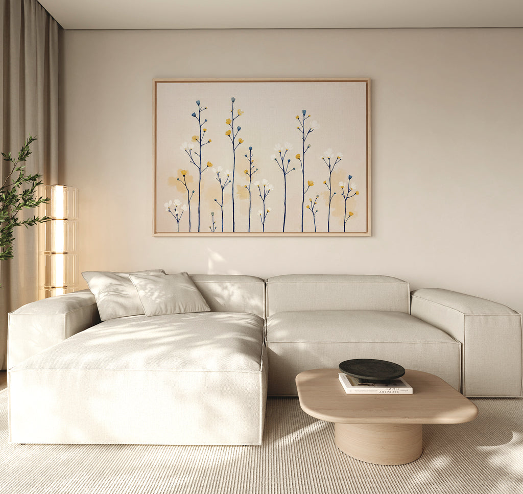 A modern living room features a light beige sectional, a low wooden coffee table, and the Meadow Symphony Canvas Art - Horizontal on the wall with minimalist blue and yellow flowers. Neutral tones and natural lighting add to the serene atmosphere.