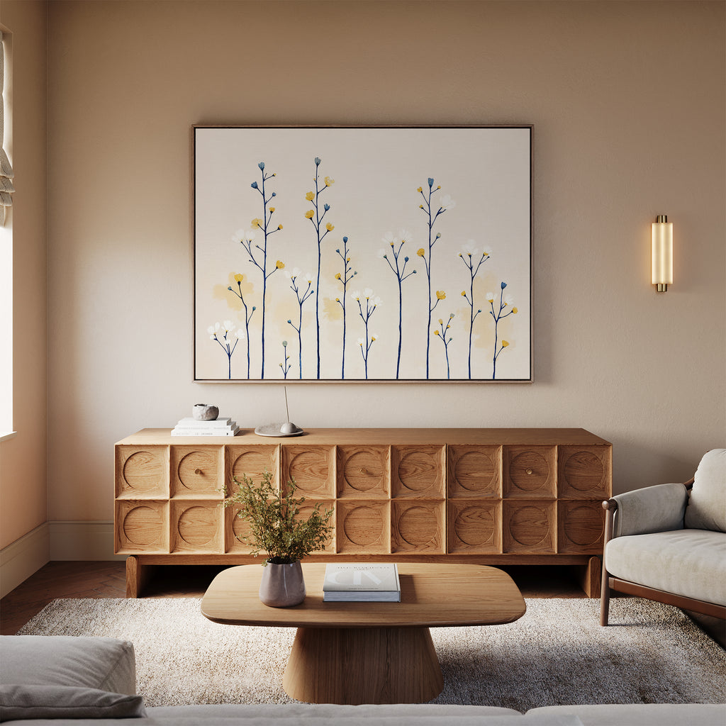 A cozy living room features a light wood coffee table, matching sideboard, beige sofa, soft rug, and the Meadow Symphony Canvas Art - Horizontal depicting minimalist branches with yellow and blue leaves. A small vase with flowers rests on the table.