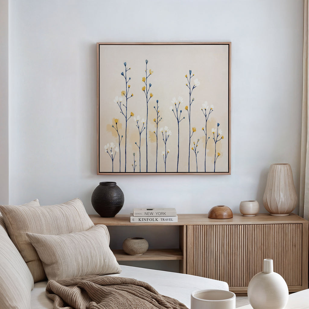 A minimalist living room features a light sofa, beige and cream cushions, a wooden sideboard with vases and books, and the Meadow Symphony Canvas Art - Square showcasing yellow and blue wildflowers for fresh spring meadow decor.