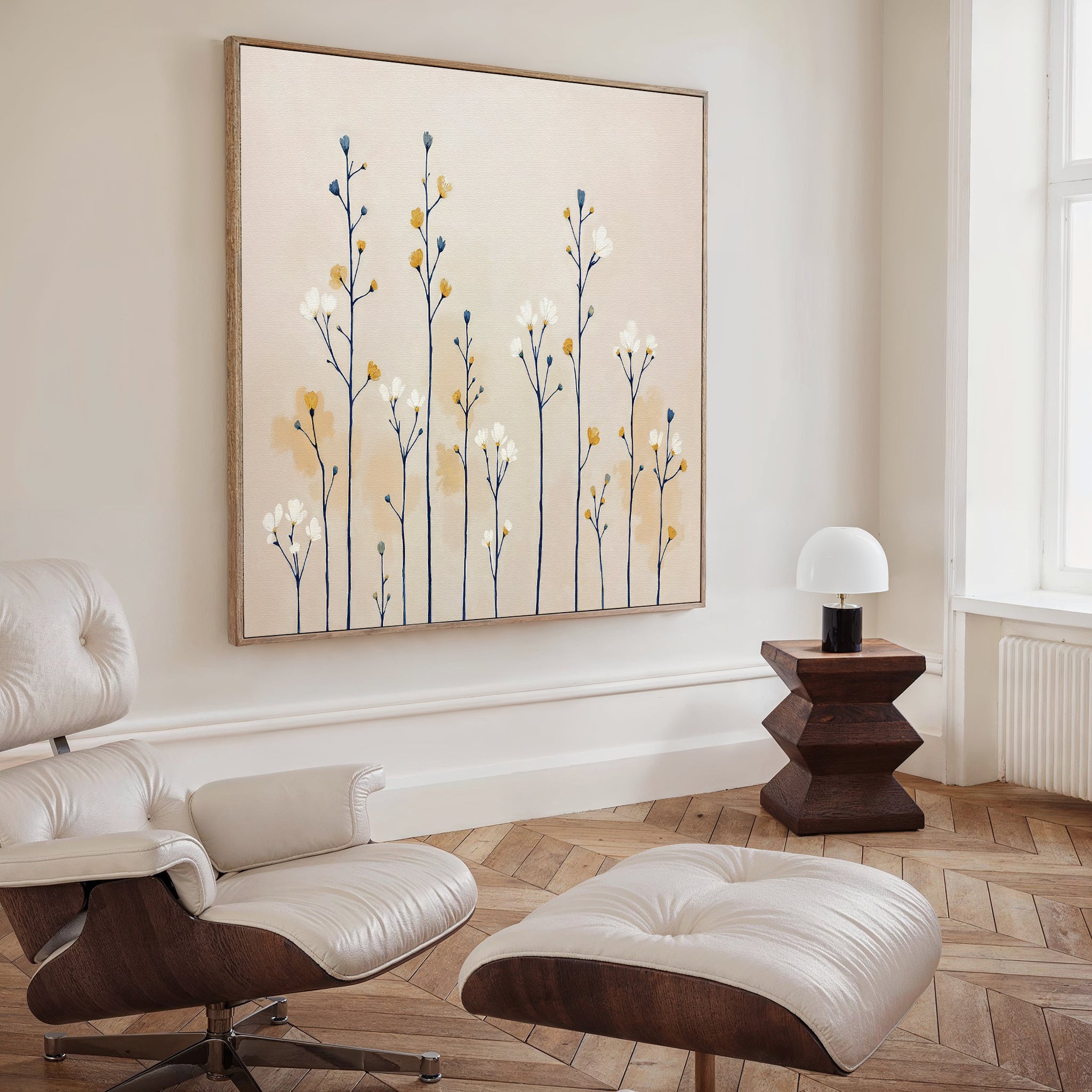 A modern, minimal living room with a white lounge chair and ottoman, geometric wooden side table with lamp, and the Meadow Symphony Canvas Art - Square featuring blue and yellow botanicals on the wall. Light wood herringbone floor.