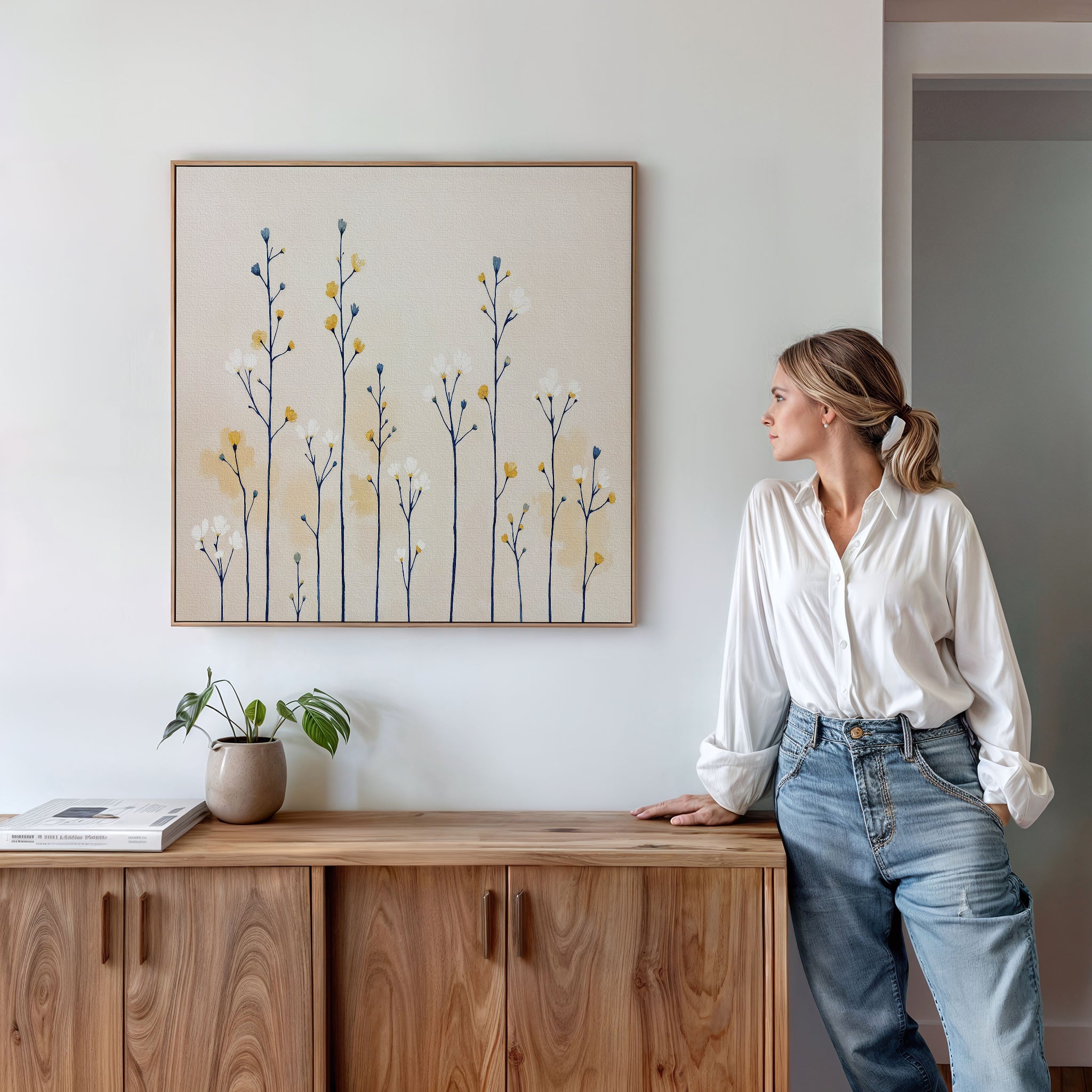 A woman in a white blouse and jeans leans on a wooden cabinet, admiring the Meadow Symphony Canvas Art - Square. A potted plant and book nearby enhance the room's calm, modern vibe.