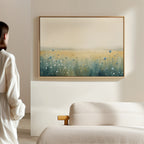A person in a white shirt stands in a bright, minimal room, admiring the Meadow Whispers Canvas Art - Horizontal—an impressionistic blue and white wildflower landscape—displayed above a beige sofa.