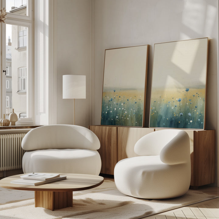 A modern living room features two white rounded armchairs, a wooden coffee table, floor lamp, and the Meadow Whispers - Set of 2 canvas prints—impressionistic landscapes—displayed on a wooden sideboard with light from tall windows.