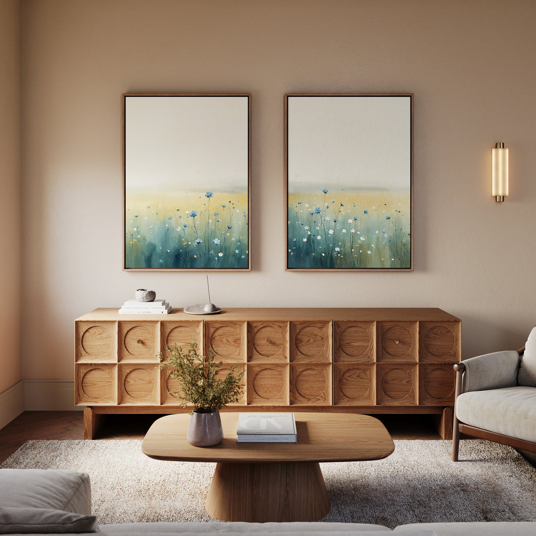 A cozy living room features a modern wooden sideboard, matching coffee table, light sofa, and two Meadow Whispers - Set of 2 landscape paintings above the sideboard. A small plant and books on the table add a tranquil touch.