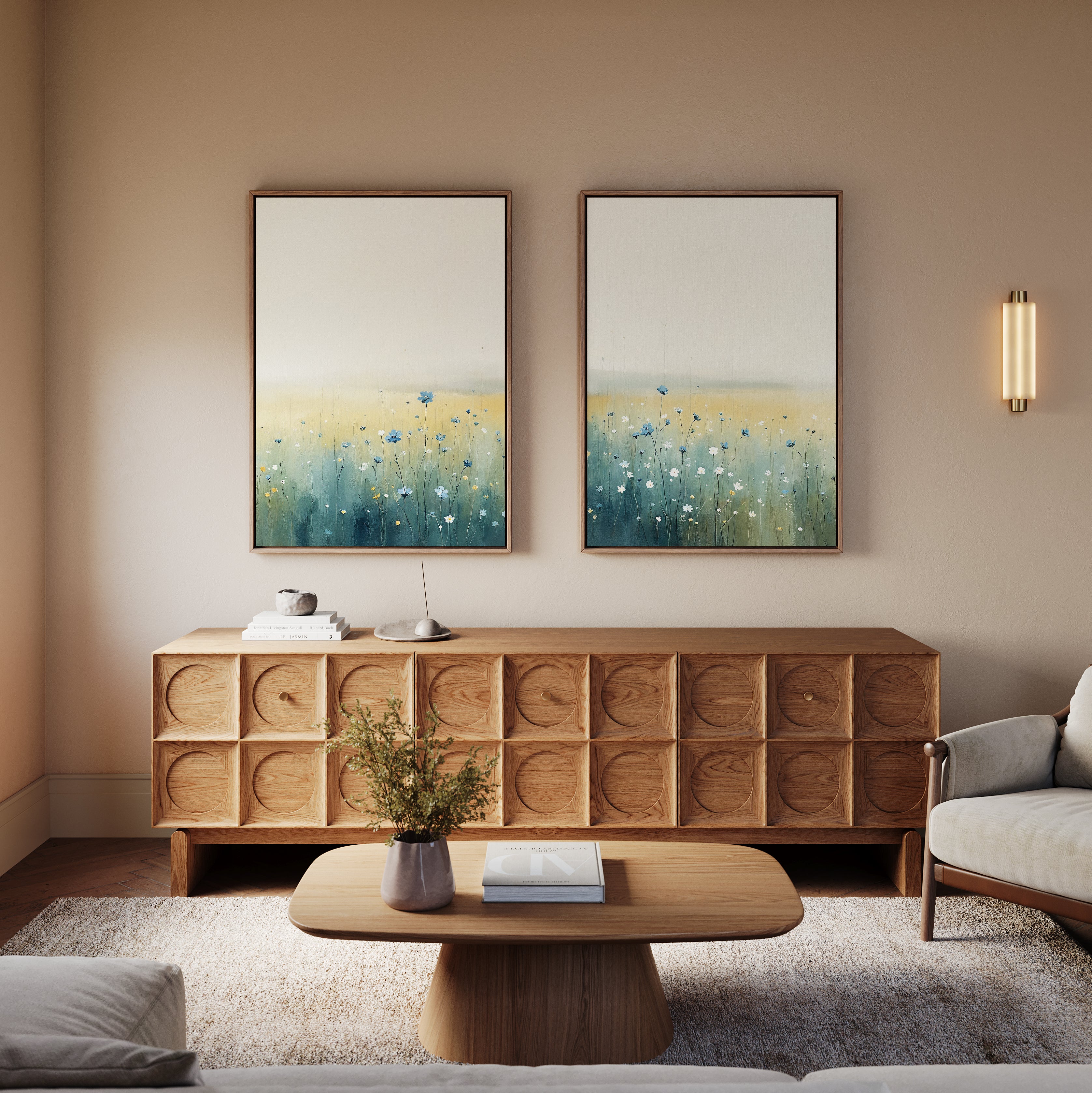 A cozy living room features a modern wooden sideboard, matching coffee table, light sofa, and two Meadow Whispers - Set of 2 landscape paintings above the sideboard. A small plant and books on the table add a tranquil touch.