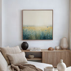 A modern, minimal living room in neutral tones features a beige sofa with cushions, a wooden sideboard with decor accents, and the Meadow Whispers Canvas Art - Square displaying impressionistic wildflowers in blue, green, and yellow on the wall.