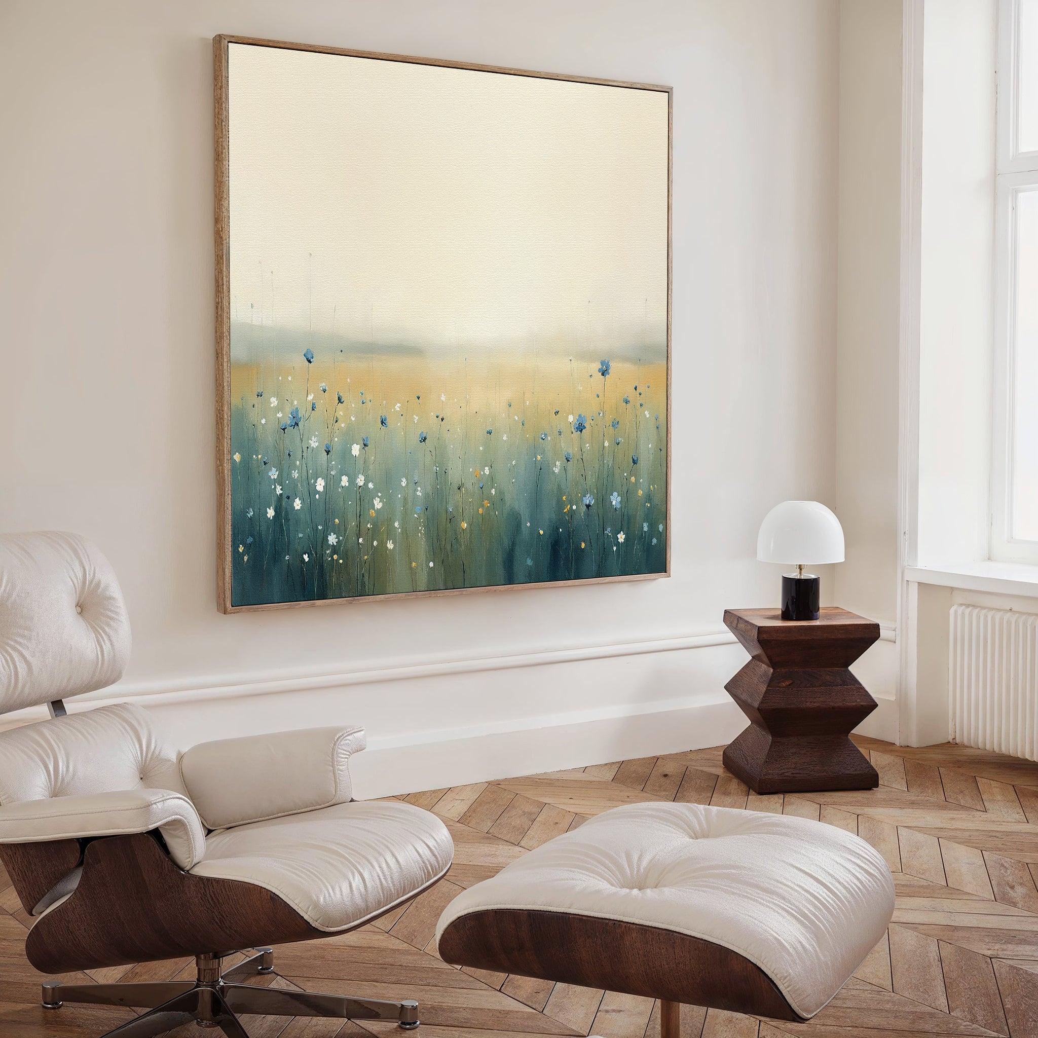 A modern living room with a white lounge chair and ottoman, a wooden side table with a small white lamp, and the Meadow Whispers Canvas Art - Square featuring blue and yellow wildflowers hanging on the wall.