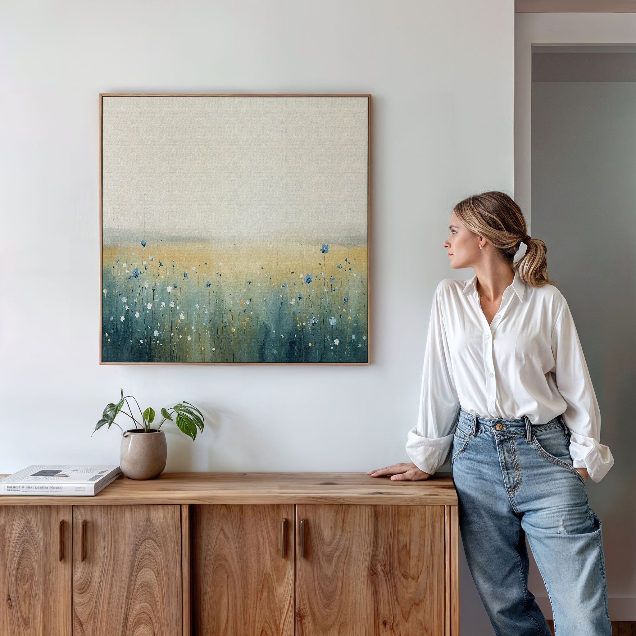 A woman in a white blouse and jeans leans on a wooden cabinet, gazing at the Meadow Whispers Canvas Art - Square, which showcases an impressionistic landscape with wildflowers and soft yellow-blue tones on the wall above her.