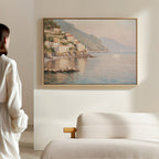 A person stands in a softly lit room, admiring the Mediterranean Dreams Canvas Art - Horizontal, which portrays a coastal village with colorful cliffside houses and calm blue water. A beige sofa in the foreground enhances the serene décor.