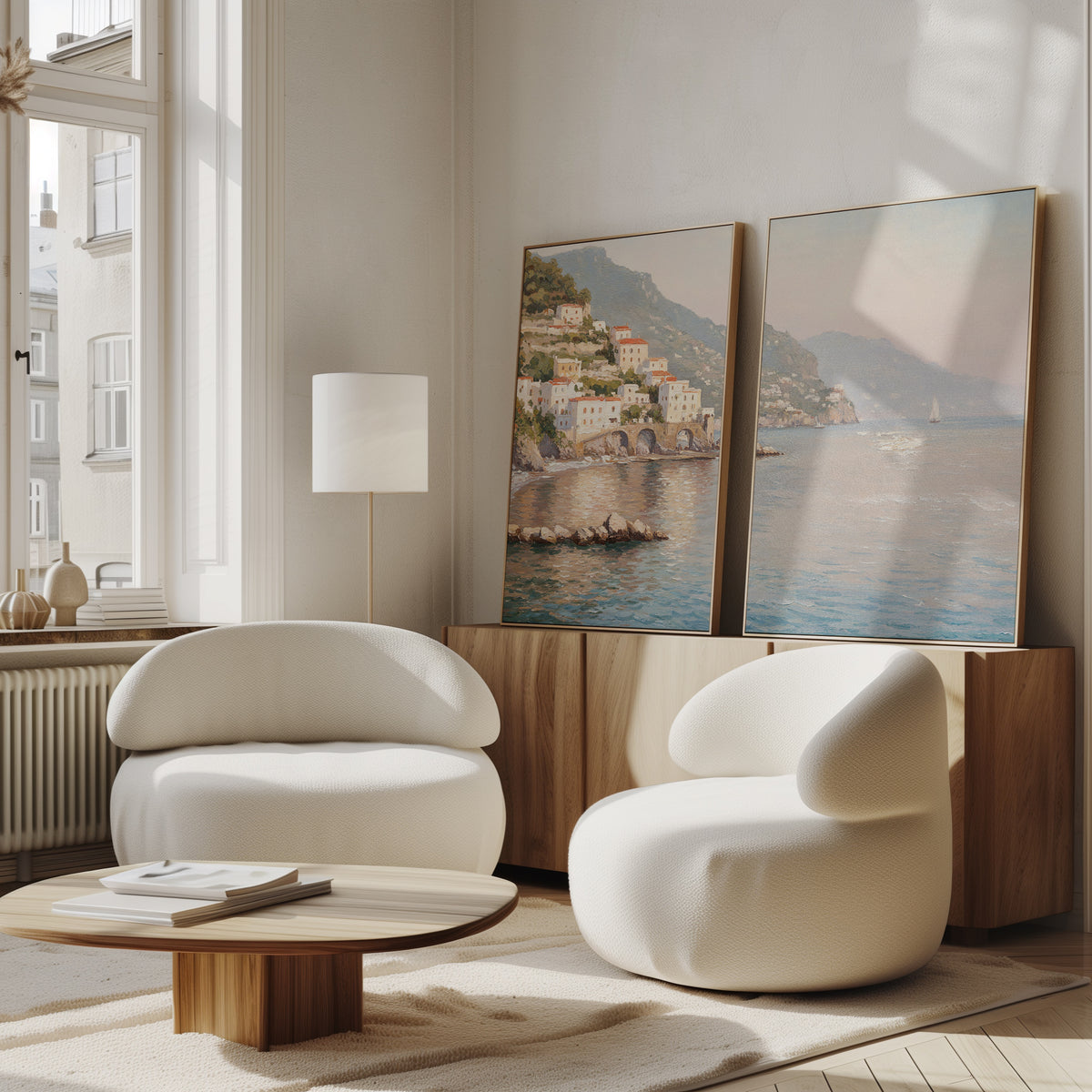 A modern living room with two curved white armchairs, a round wooden coffee table, and serene decor featuring the "Mediterranean Dreams - Set of 2" coastal village wall art leaning against a wooden sideboard by a large window.