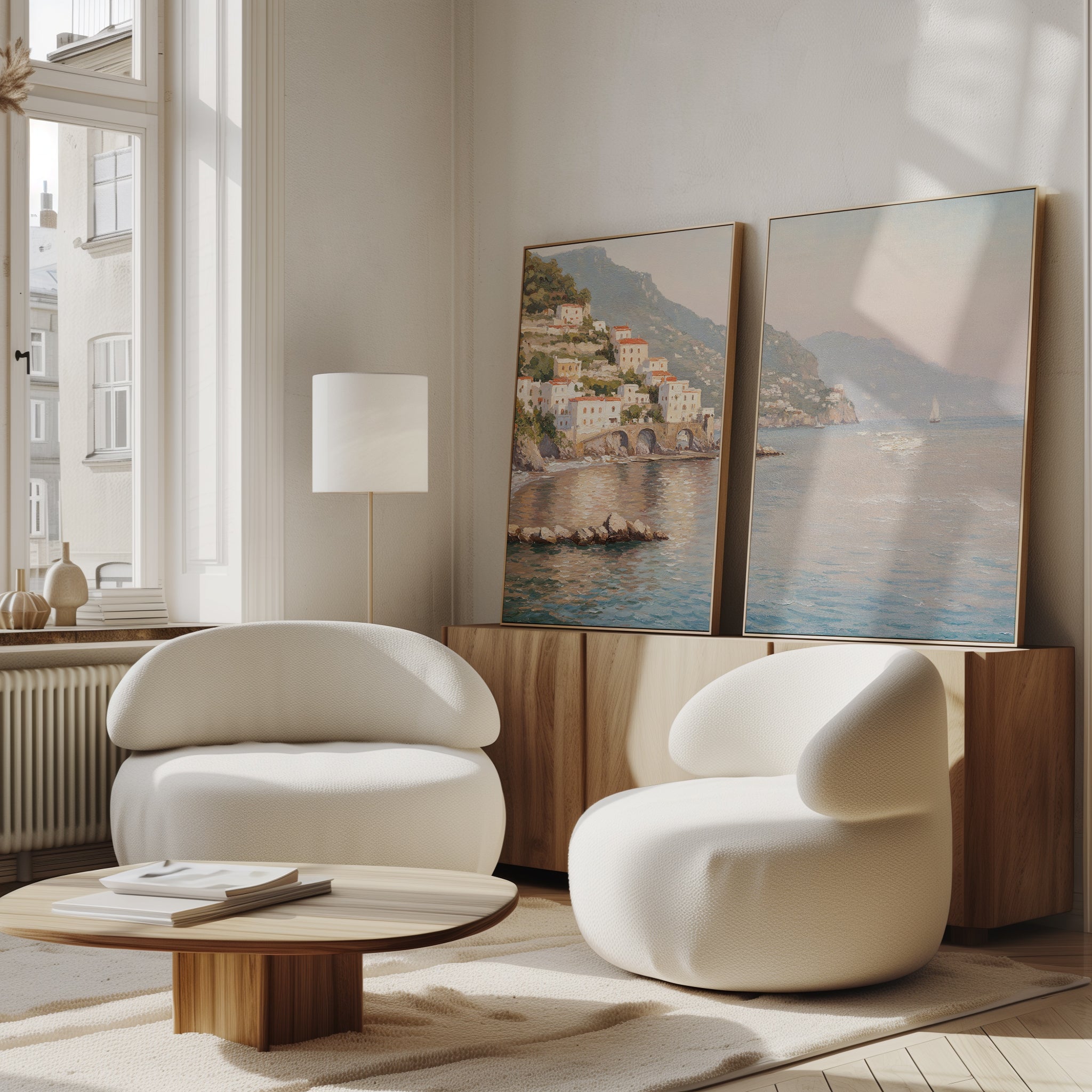 A modern living room with two curved white armchairs, a round wooden coffee table, and serene decor featuring the "Mediterranean Dreams - Set of 2" coastal village wall art leaning against a wooden sideboard by a large window.