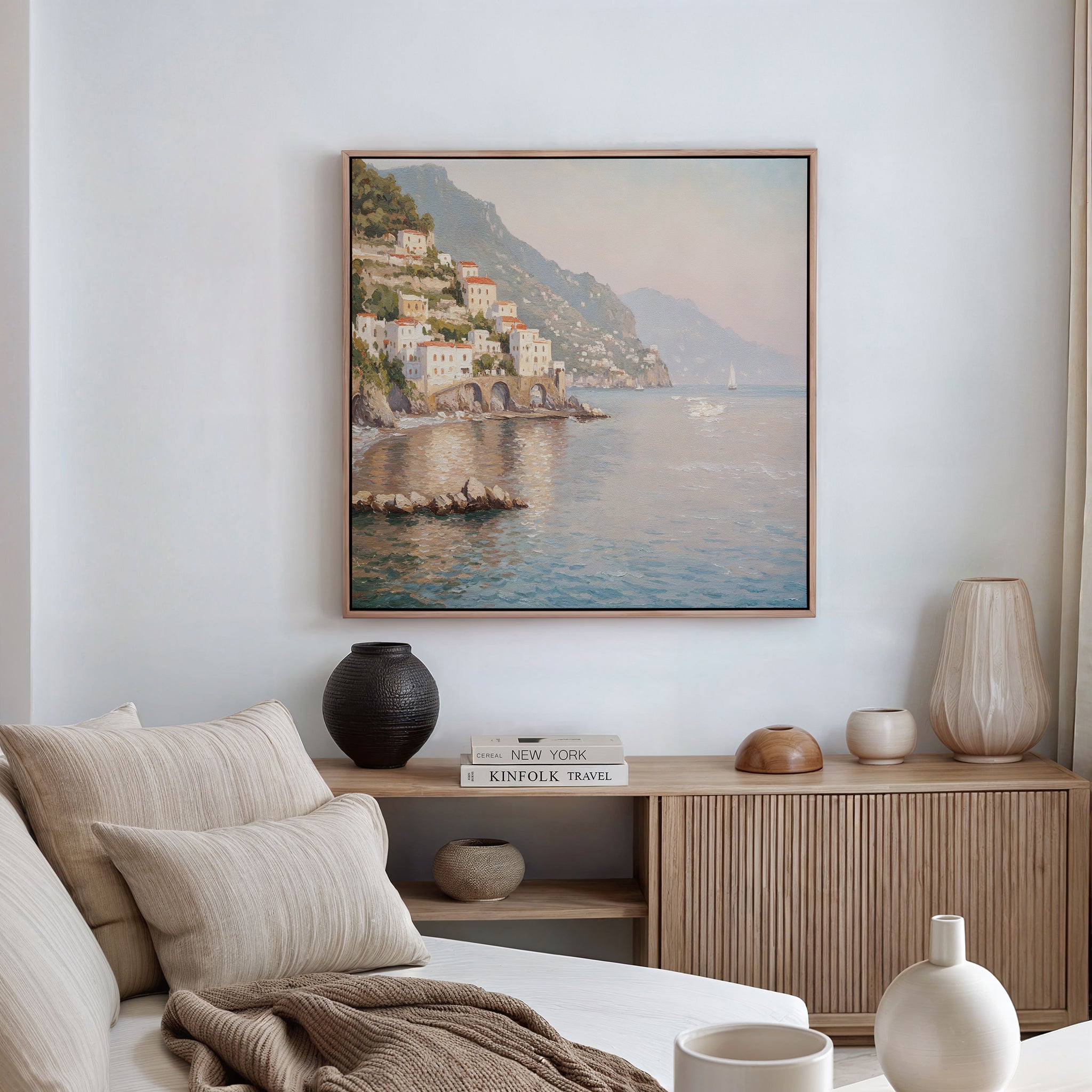 A cozy, neutral-toned living space features a beige sofa, wooden sideboard with vases and books, and serene decor highlighted by the Mediterranean Dreams Canvas Art - Square depicting a coastal village on the white wall.