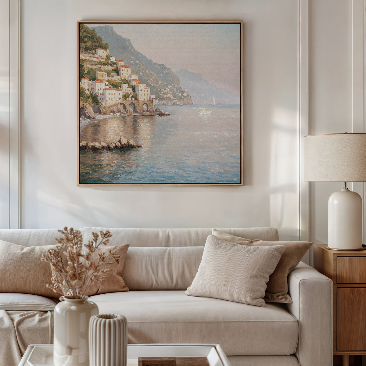 A cozy living room with a beige sofa, cream cushions, a wooden side table, and dried flowers. Above the sofa hangs the Mediterranean Dreams Canvas Art - Square, featuring a coastal village for serene home decor.