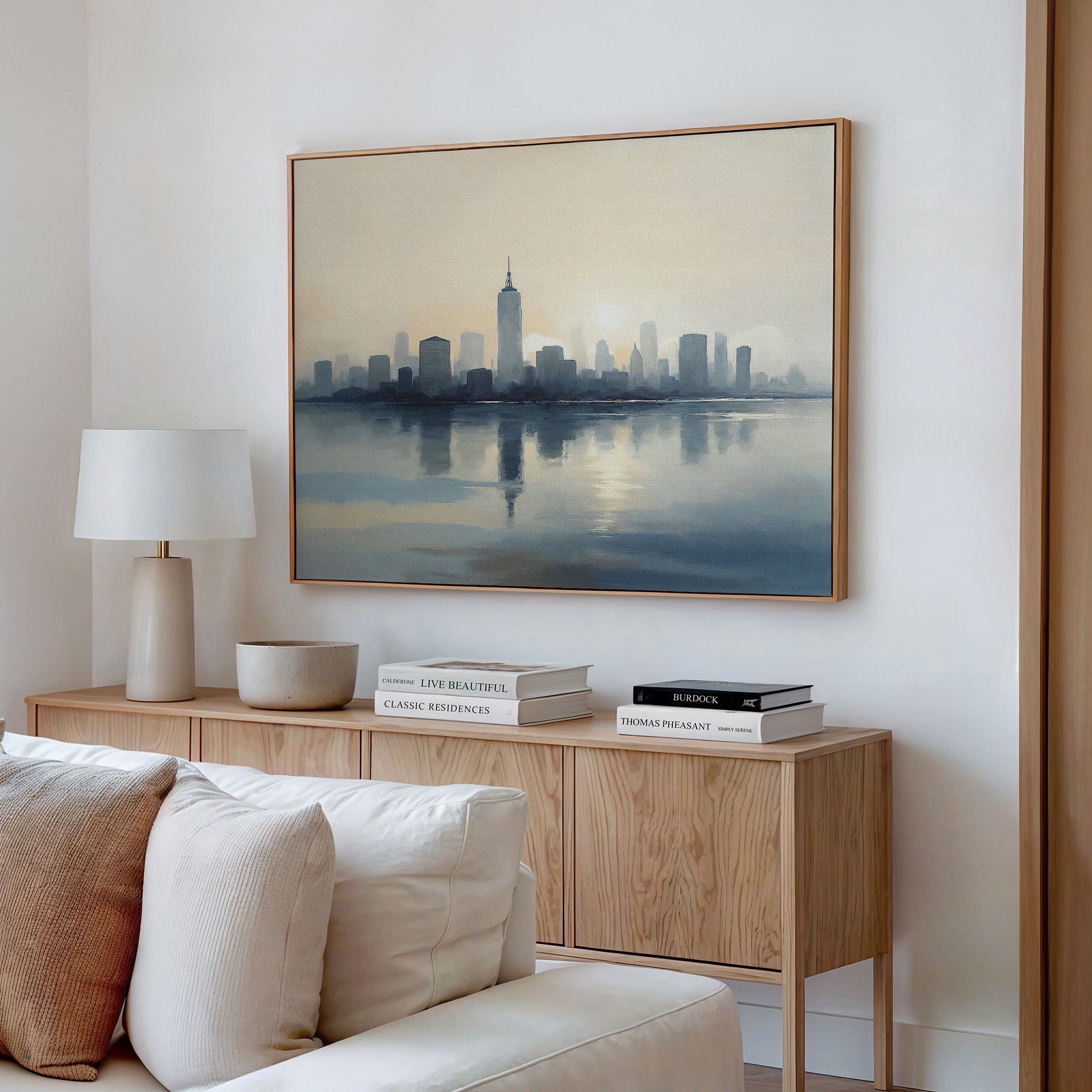 A modern living room features a beige sofa, light wood console table, white lamp, decorative bowls, stacked books, and urban tranquility brought by the Metropolitan Dawn Canvas Art - Horizontal depicting a city skyline reflected on water.