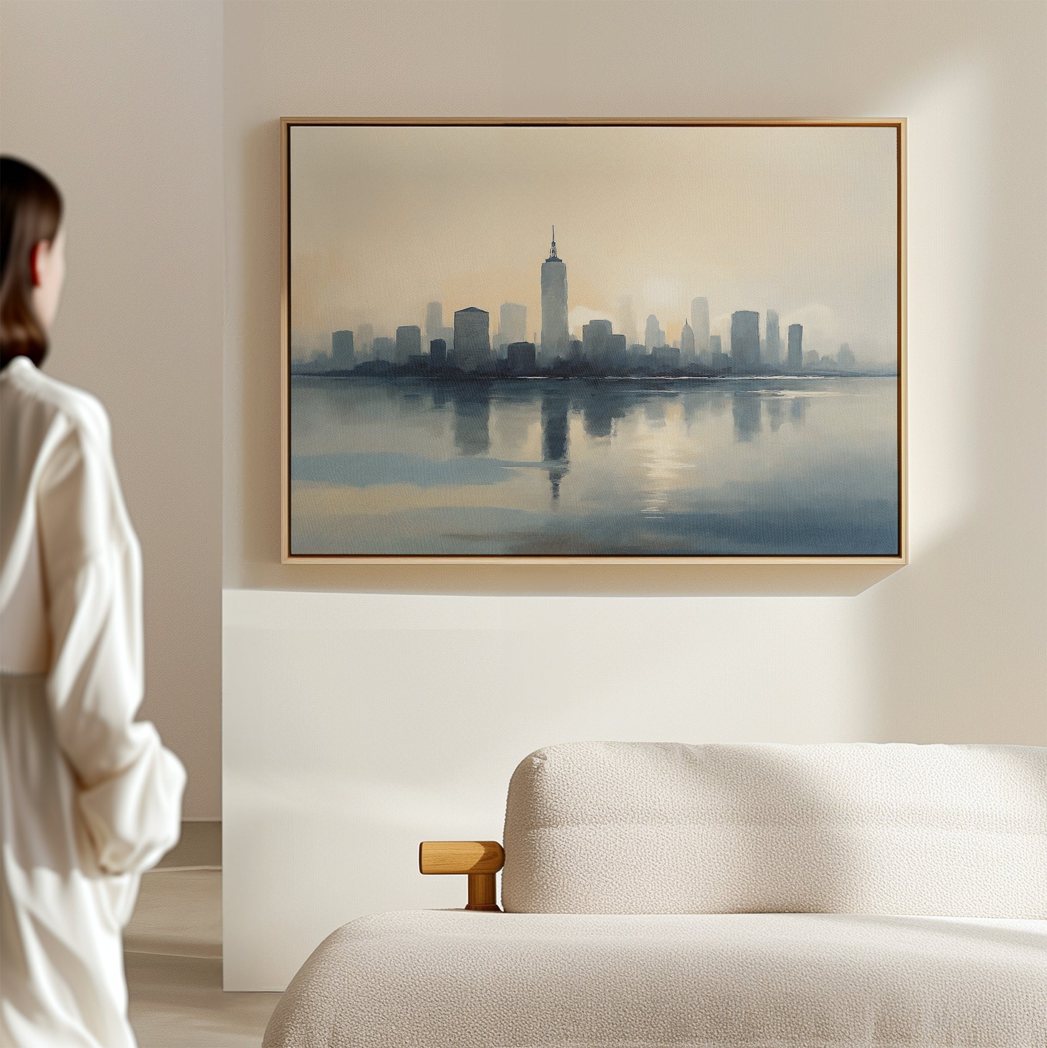 A woman stands in a minimal, light-filled room, gazing at the Metropolitan Dawn Canvas Art - Horizontal, a serene city skyline above calm water, displayed above a beige sofa.