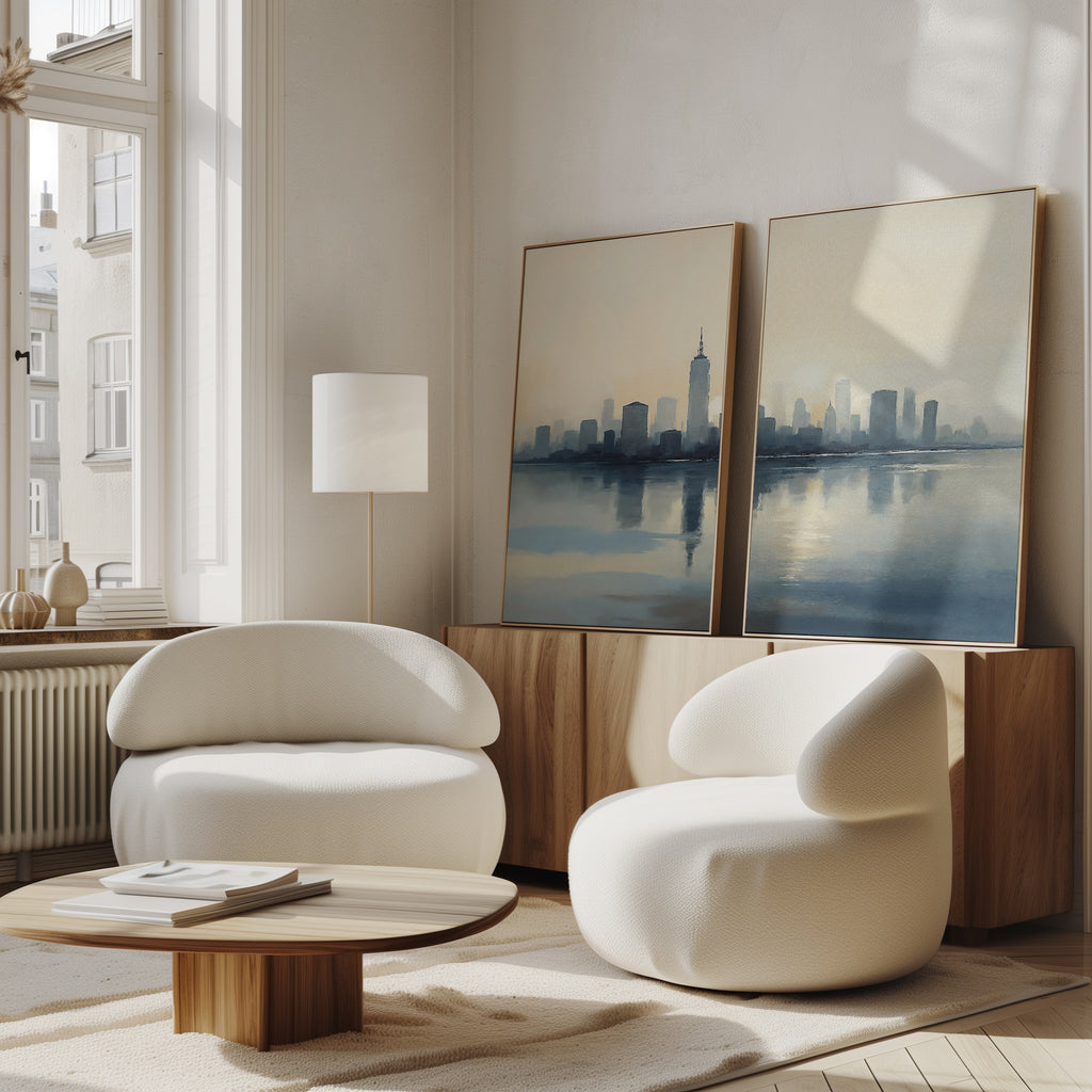 A modern living room features two white, rounded armchairs, a low wooden coffee table, and the Metropolitan Dawn - Set of 2 canvas prints on a sideboard. Large windows fill the cozy, minimalist space with calming natural light and urban tranquility.