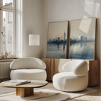 A modern living room features two white, rounded armchairs, a low wooden coffee table, and the Metropolitan Dawn - Set of 2 canvas prints on a sideboard. Large windows fill the cozy, minimalist space with calming natural light and urban tranquility.