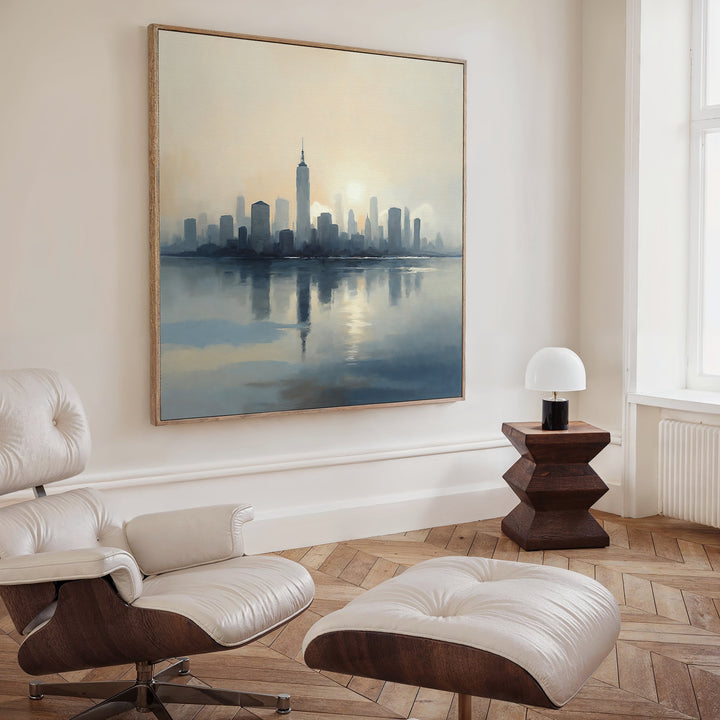 A modern living room with a white leather lounge chair and ottoman, wooden side table with lamp, and the Metropolitan Dawn Canvas Art - Square featuring a vivid city skyline over water. Light pours in from tall windows, emphasizing the elegant urban vibe.