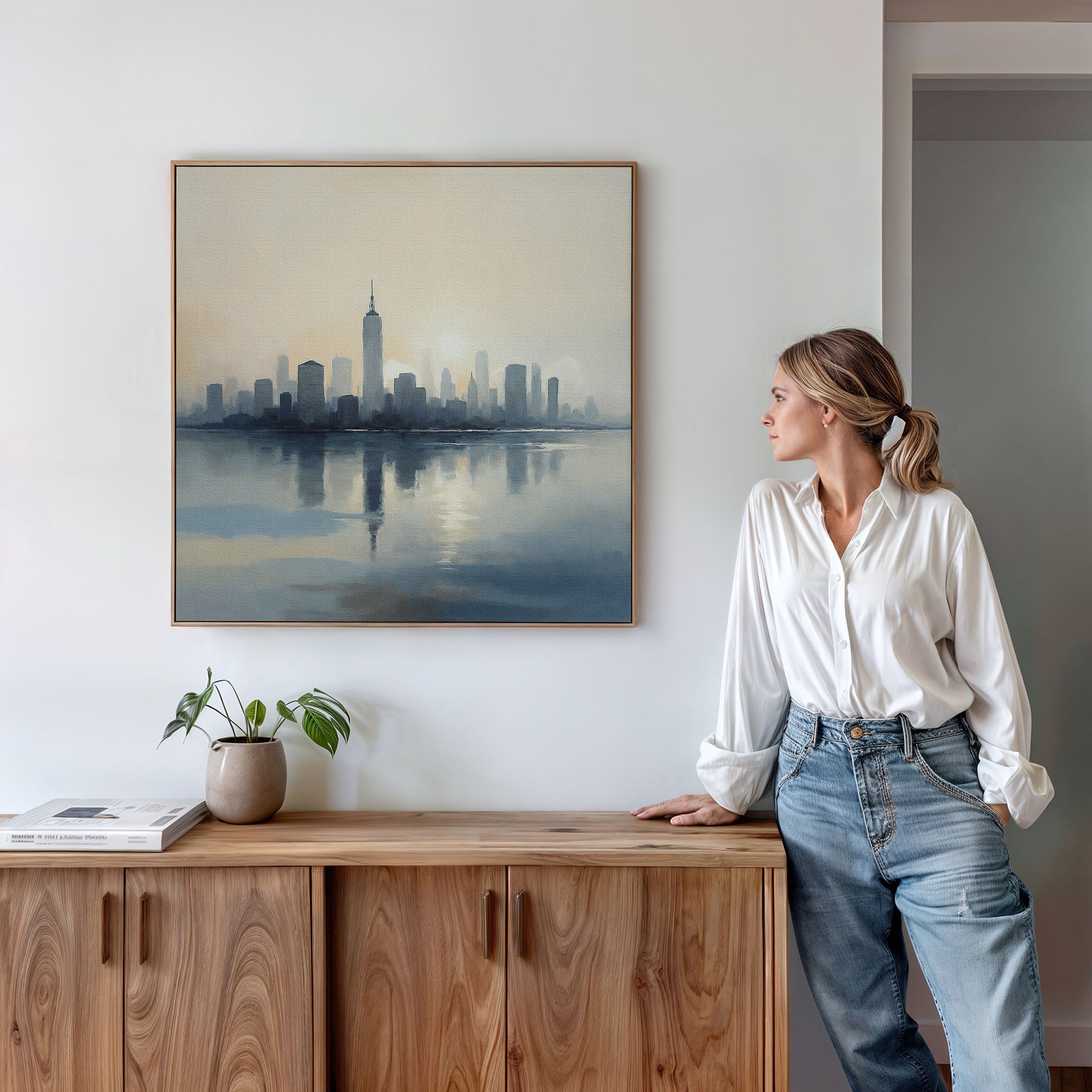 A woman in a white blouse and jeans stands by a wooden cabinet, admiring the Metropolitan Dawn Canvas Art - Square. A potted plant and books on the cabinet add charm to the urban wall art displayed on the white wall.