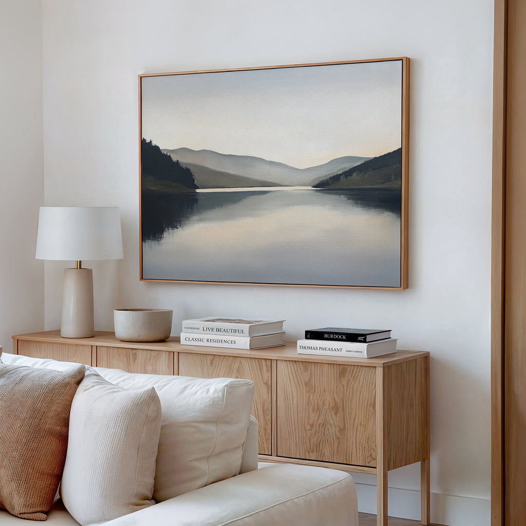 A minimalist living room features a light wood sideboard, white lamp, and decorative bowl. Above, the Mirror Valley Canvas Art - Horizontal adds serene beauty with its calm natural palette.