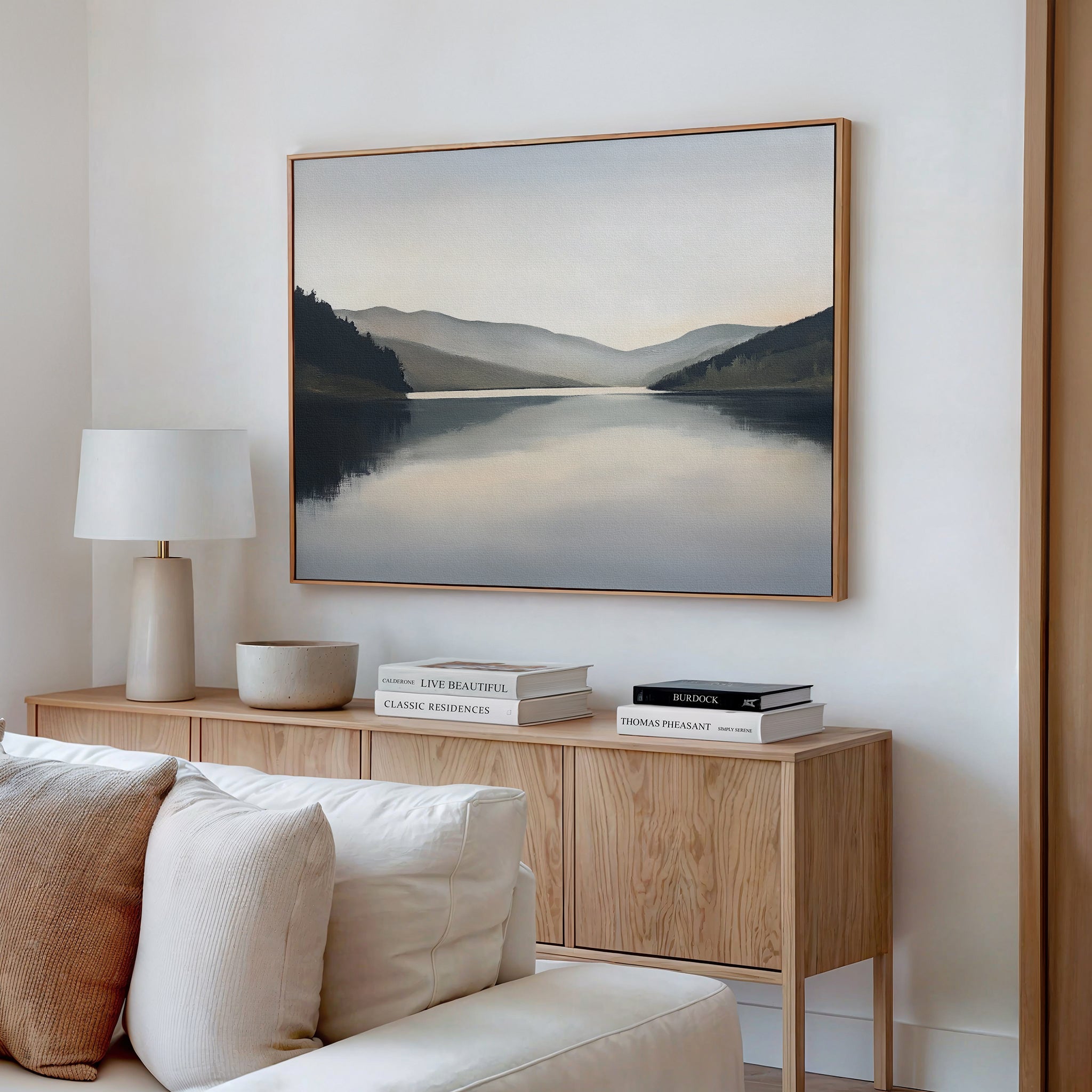 A minimalist living room features a light wood sideboard, white lamp, and decorative bowl. Above, the Mirror Valley Canvas Art - Horizontal adds serene beauty with its calm natural palette.
